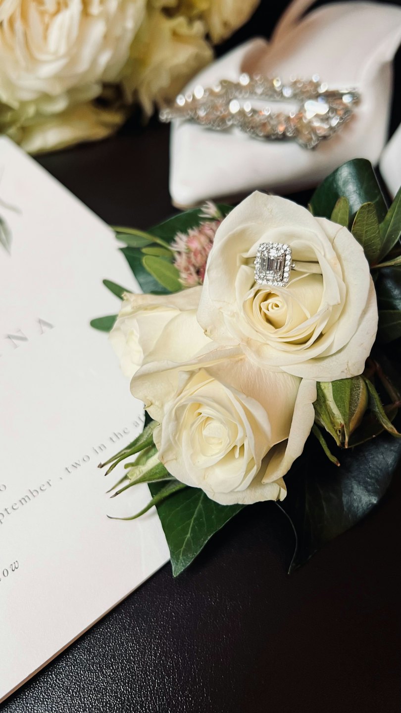 Anello di fidanzamento su rose bianche accanto a un invito di nozze.